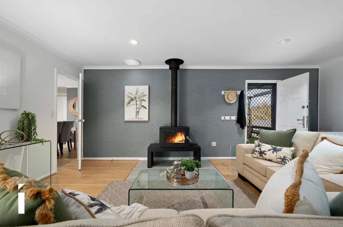 A living area is showcased with a modern design, featuring a black wood stove as a focal point. A neutral-colored couch is complemented by decorative pillows. A glass coffee table rests on a light wooden floor, with greenery and a decorative bowl enhancing the space's appeal.