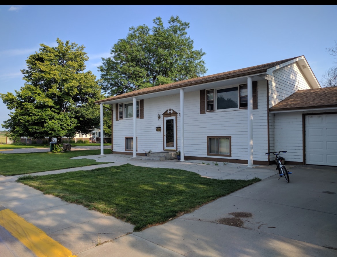 A two-story house is shown with a light-colored exterior, featuring a front porch supported by white columns. The driveway leads to a garage door on the right, and a bicycle is parked nearby. Lush green grass complements the well-maintained landscape.