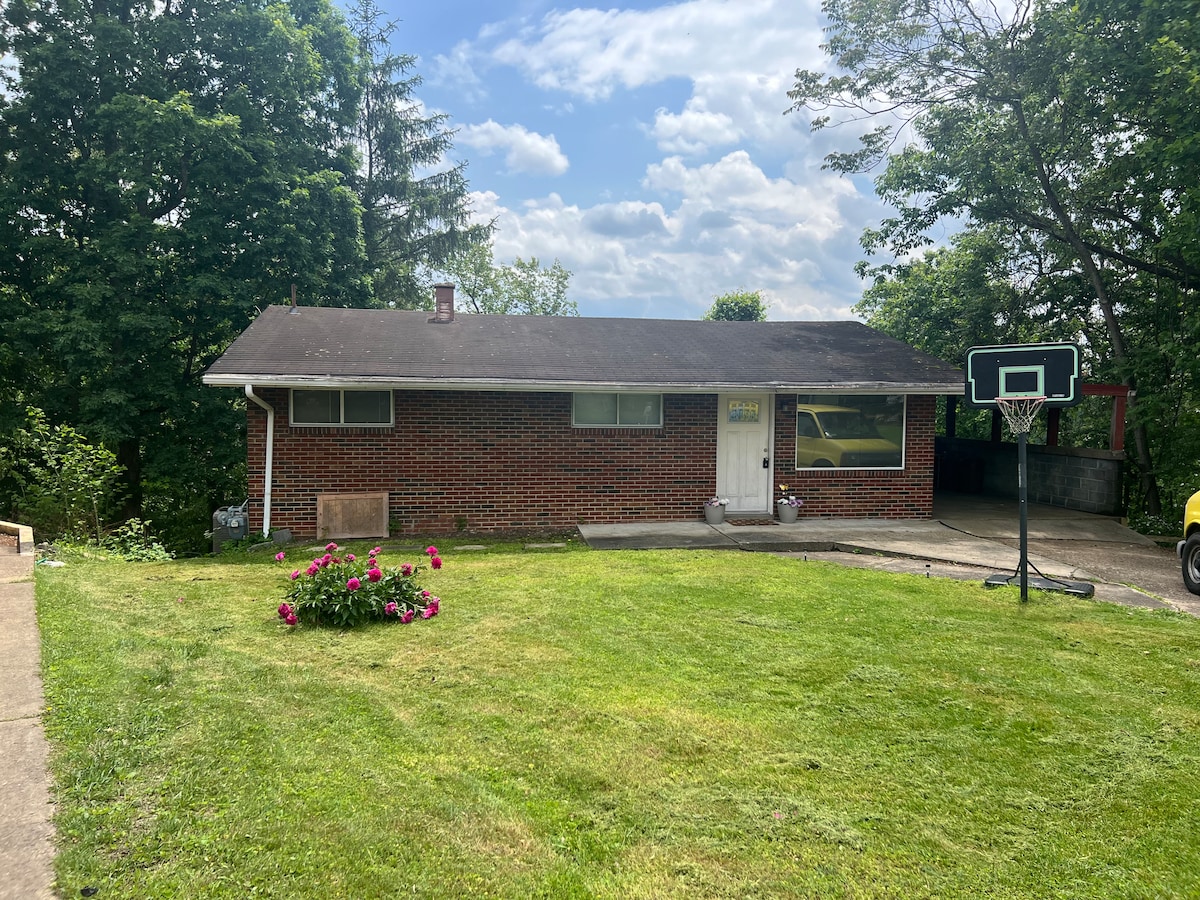 A single-story brick house is set on a grassy lawn adorned with pink flowers. A basketball hoop is visible on the driveway, and a tree line provides a natural backdrop. The sky is partly cloudy, allowing soft light to illuminate the scene.