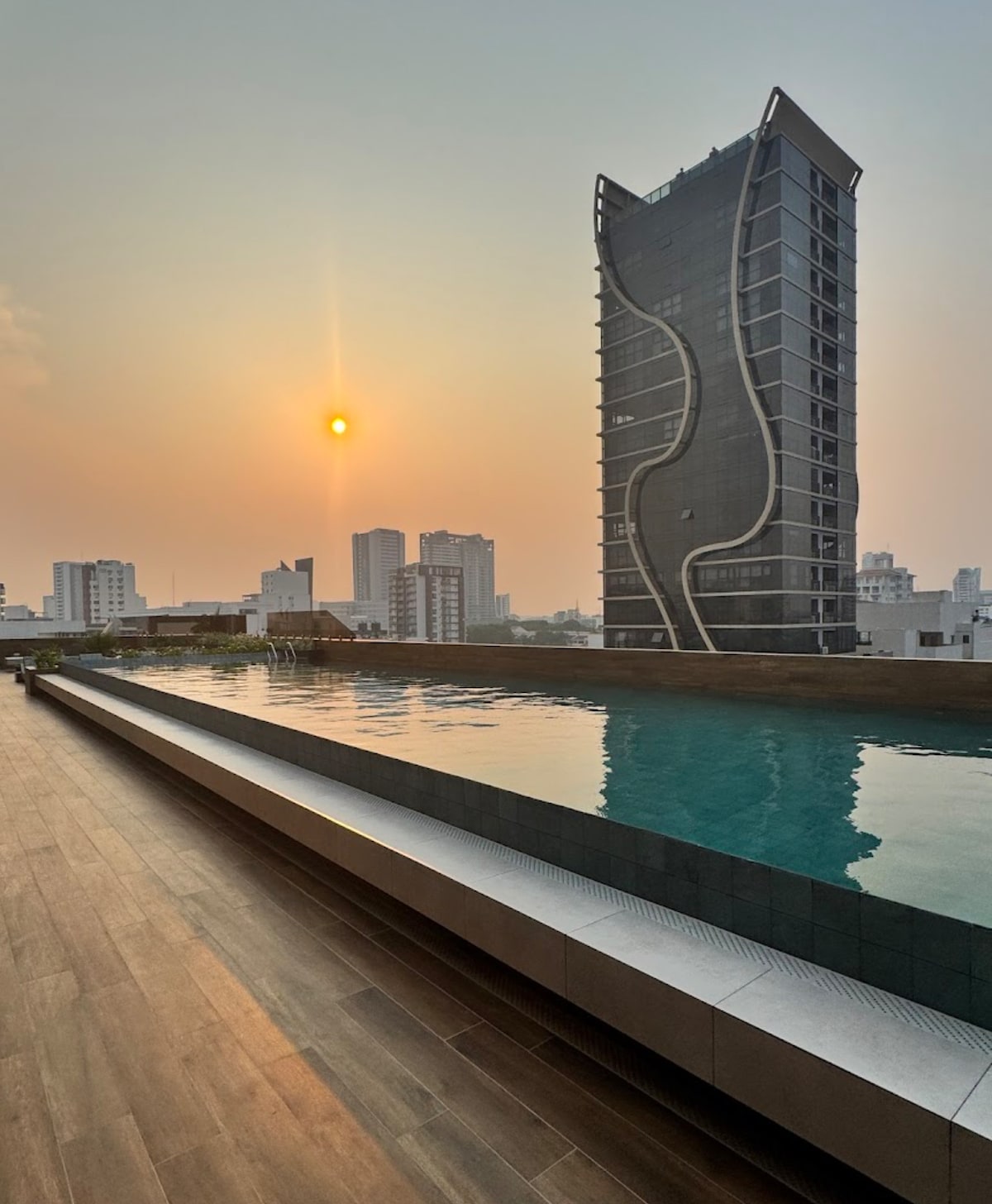 A rooftop swimming pool reflects the soft light of the setting sun, positioned alongside sleek modern architecture. The view captures the urban skyline of the city, with various high-rise buildings that complement the contemporary design of the surrounding environment.