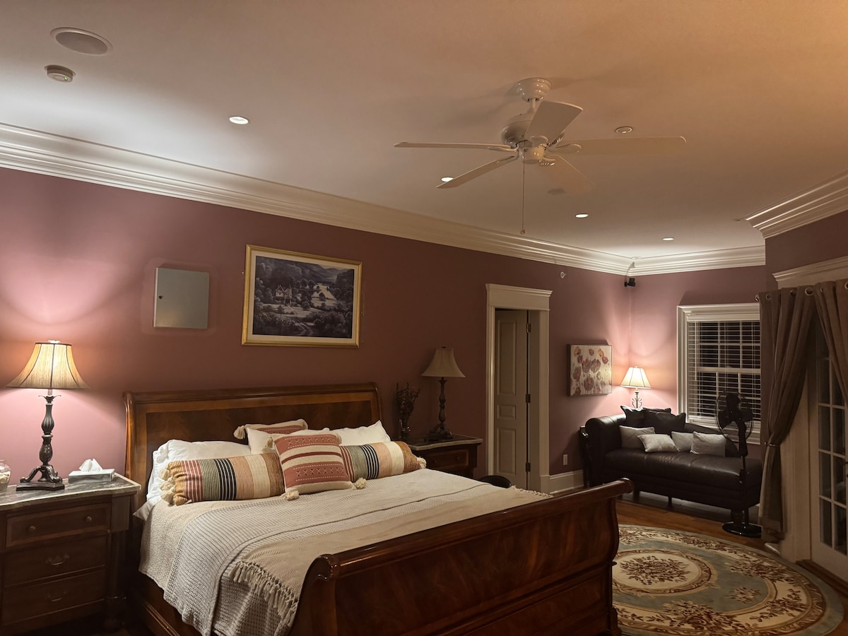 A spacious bedroom is featured with a wooden bed frame draped in a light blanket and accented by colorful pillows. A pair of bedside lamps provide soft lighting. In the corner, a dark couch offers additional seating next to a window dressed with light curtains.