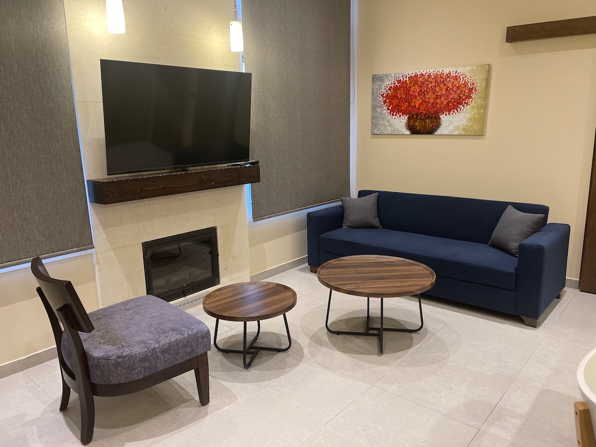 A modern living area is presented, featuring a dark blue sofa facing a wall-mounted television. Two round coffee tables with wooden surfaces are positioned in front of the seating. A stylish chair complements the space, alongside a fireplace highlighted by neutral tiles.