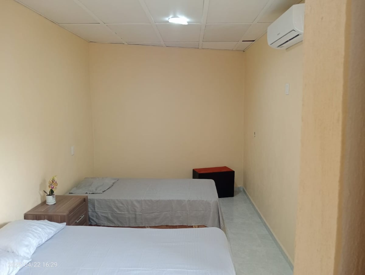 A simple sleeping area is presented, featuring two unadorned beds with light bedding. A small nightstand with a flower is positioned beside one bed, and a storage ottoman is visible in the corner. Soft lighting is provided from a ceiling fixture.