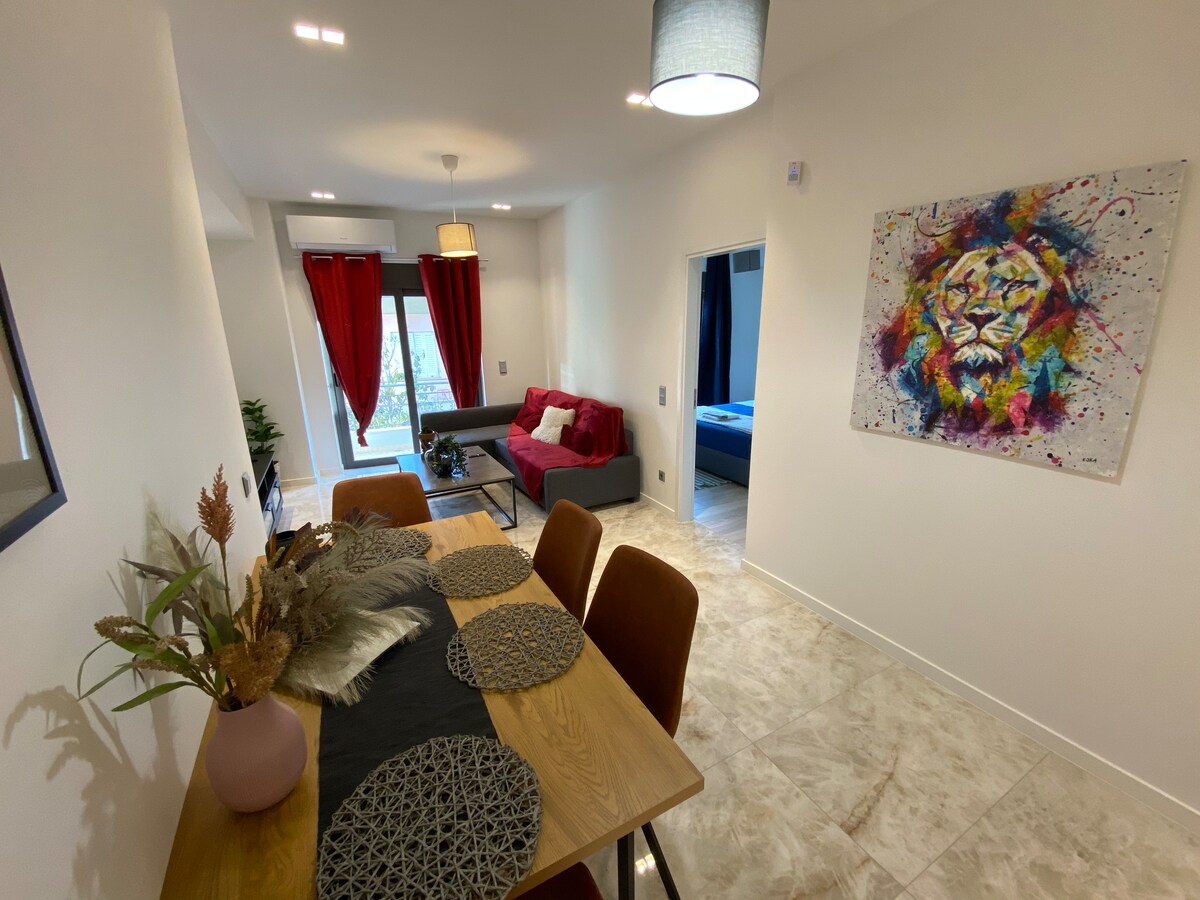 An open-concept living area features a dining table set for six, complemented by decorative placemats and a centerpiece. A cozy sofa is situated nearby, accented by vibrant red curtains framing the windows, and colorful artwork adds character to the light-filled space.
