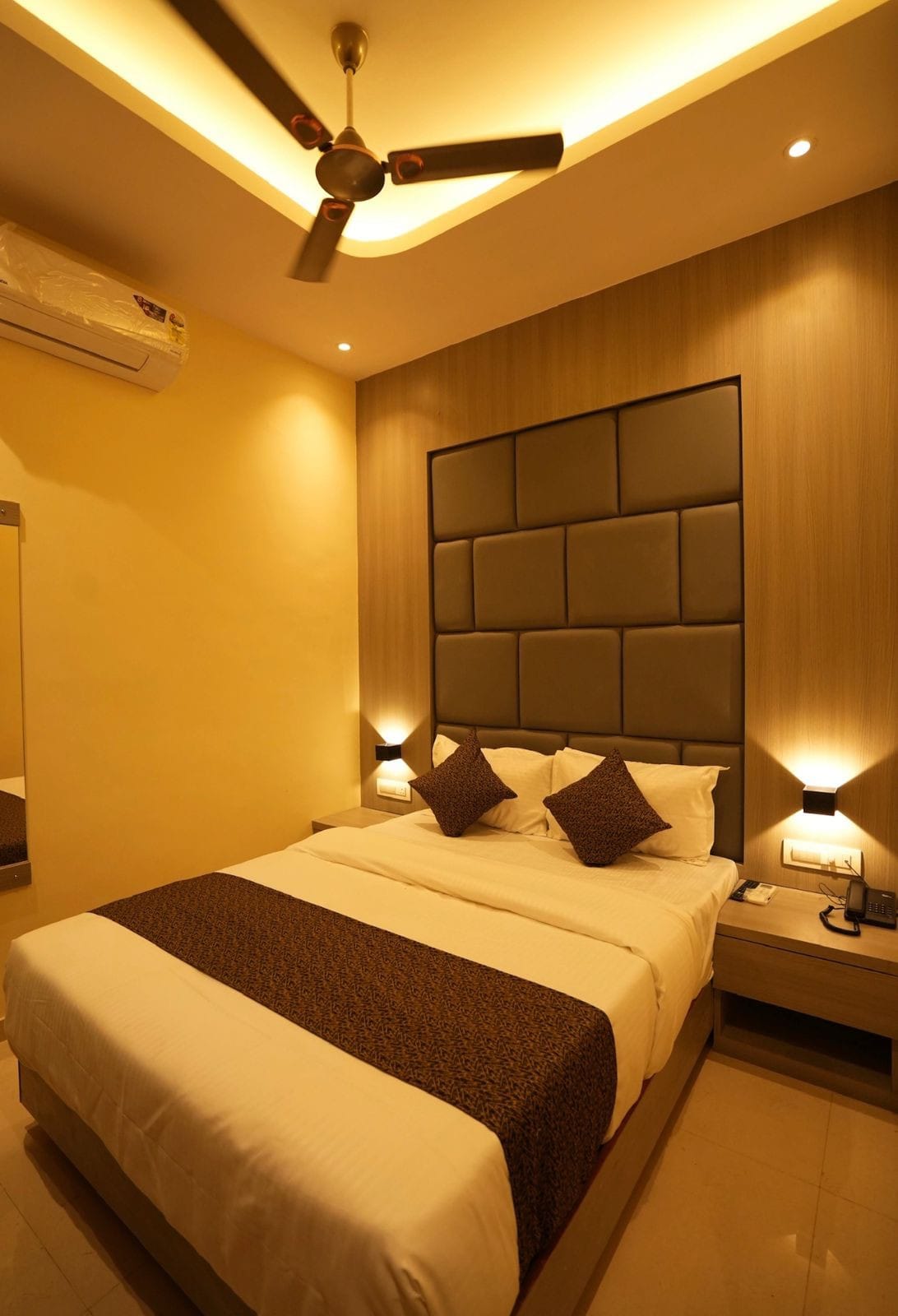 A cozy bedroom features a spacious bed adorned with decorative pillows. Soft lighting is provided by bedside lamps, and a mirror reflects the space. The ceiling is equipped with a fan, and modern wall finishes create a welcoming atmosphere.