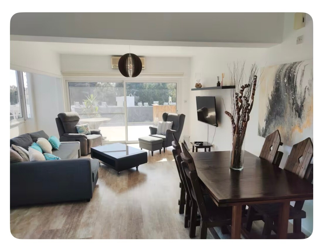 A modern living space features a large dining table surrounded by wooden chairs, complemented by a spacious sofa and an armchair. Natural light enters through expansive windows, offering views of an outdoor area. A wall-mounted television is positioned opposite the seating area.