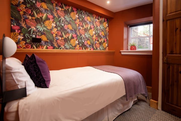 Este acogedor dormitorio cuenta con una cama individual rodeada de paredes de color naranja intenso. Una pared decorativa está cubierta con un caprichoso papel pintado tropical/botánico. La habitación, que incluye un armario, ofrece un estilo vibrante y ecléctico.