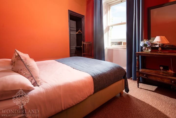 Este llamativo dormitorio cuenta con una vibrante pared naranja que contrasta dramáticamente con las cortinas y la ropa de cama de color turquesa. Contiene una cómoda cama doble y muebles clásicos de madera. La habitación ofrece vistas al mar y a la ciudad