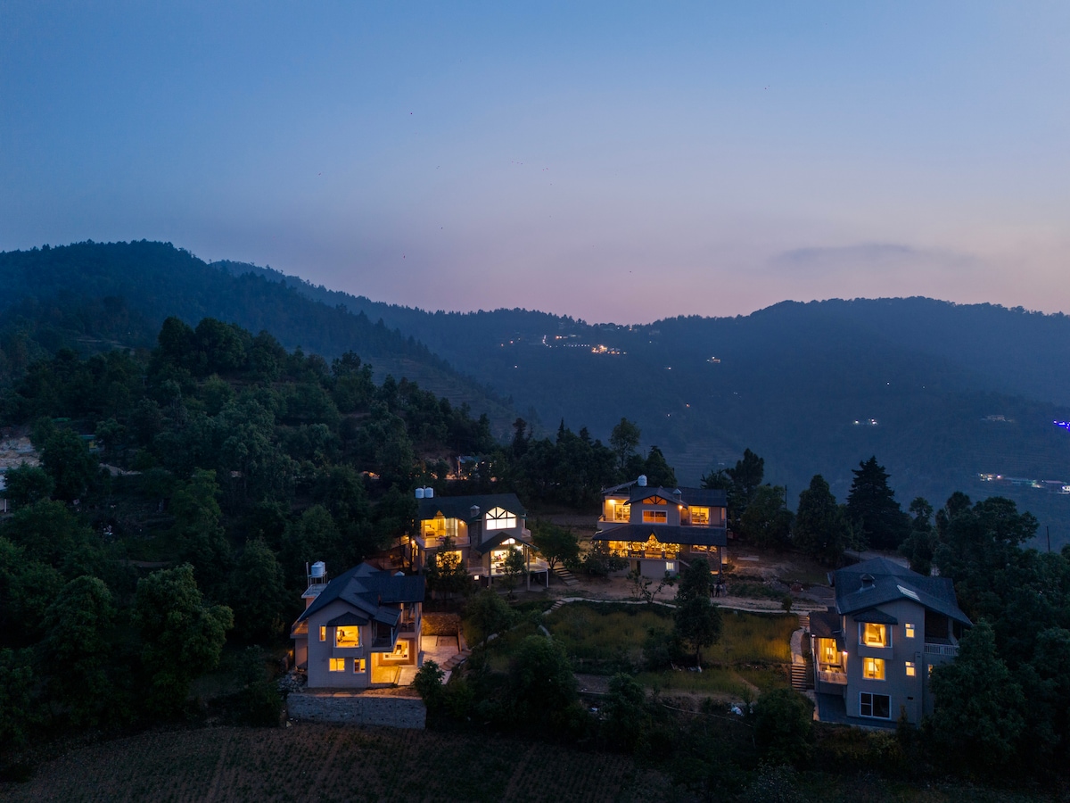 An aerial view captures multiple villas nestled among lush greenery, illuminated softly at dusk. The surrounding hills create a serene backdrop, while warm lights emanate from the windows, hinting at welcoming interiors. The well-maintained outdoor space is visible, enhancing the sense of tranquility.