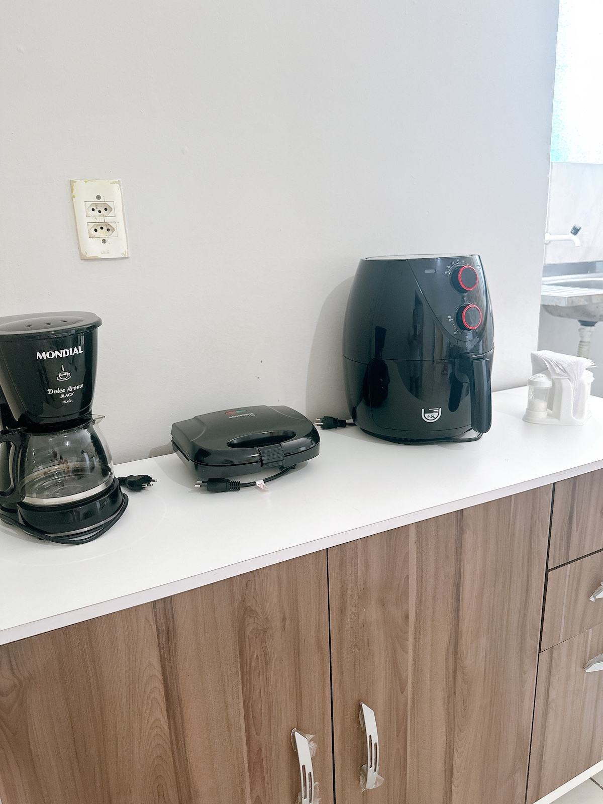 A kitchen counter is equipped with a coffee maker, a sandwich press, and an air fryer, all arranged neatly along a white surface. The cabinetry below features a warm wood finish, complementing the clean and functional design of the space.