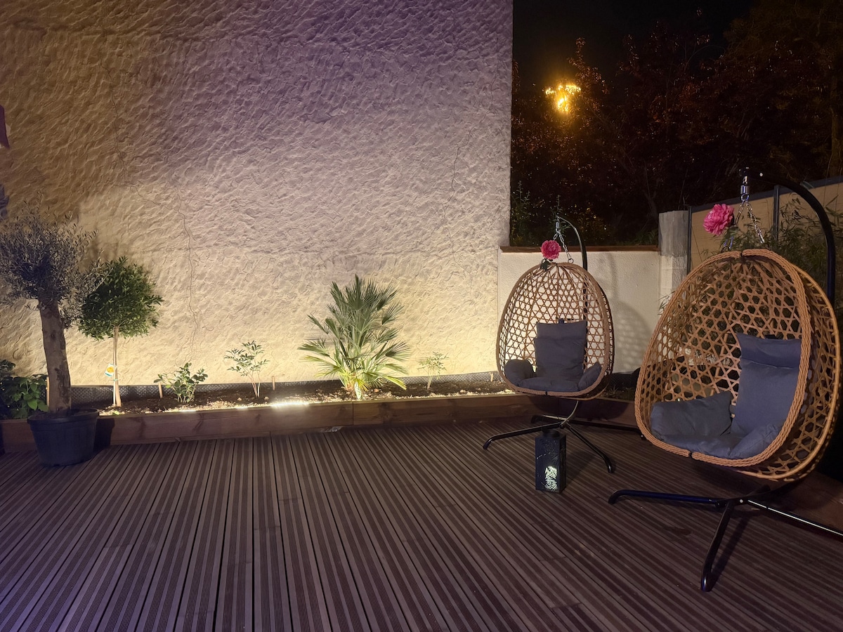 A spacious terrace features two hanging chairs with comfortable cushions, surrounded by ambient lighting. Potted plants, including an olive tree and a palm, are positioned around the area, creating a tranquil outdoor space, perfect for relaxation.