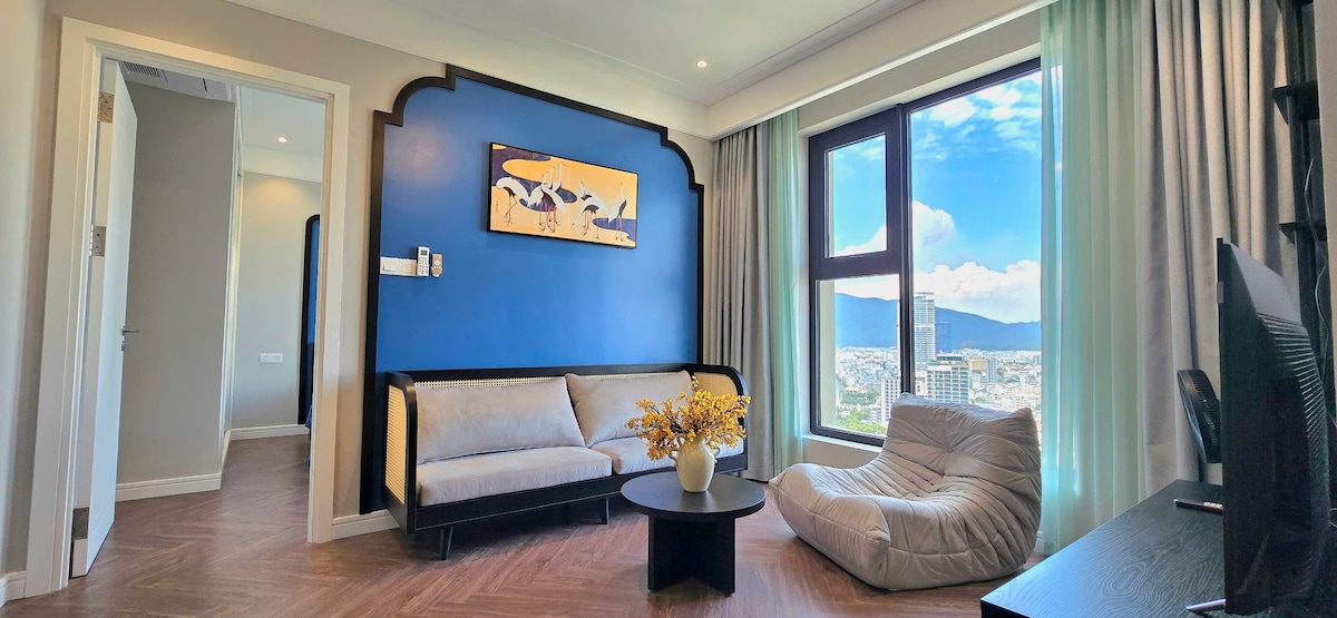 A modern living space features a stylish blue accent wall. A light-colored sofa and bean bag surround a circular black coffee table, while large windows allow natural light and offer a view of the city skyline and mountains beyond.
