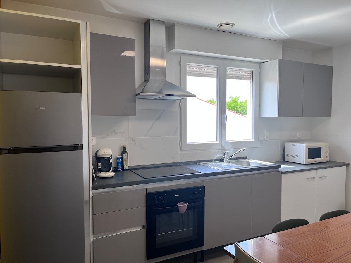 The kitchen features modern cabinetry in light grey tones, equipped with essential appliances including a refrigerator, microwave, and coffee machine. A sink is positioned between the stovetop and oven, with natural light filtering through the adjacent windows, creating a functional cooking space.