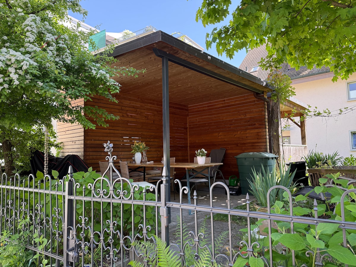 A covered outdoor seating area made of wooden panels is surrounded by lush greenery. Potted plants are positioned on a low table, creating an inviting space for relaxation. A decorative wrought iron fence enhances the garden's charm, providing a visual boundary to the area.