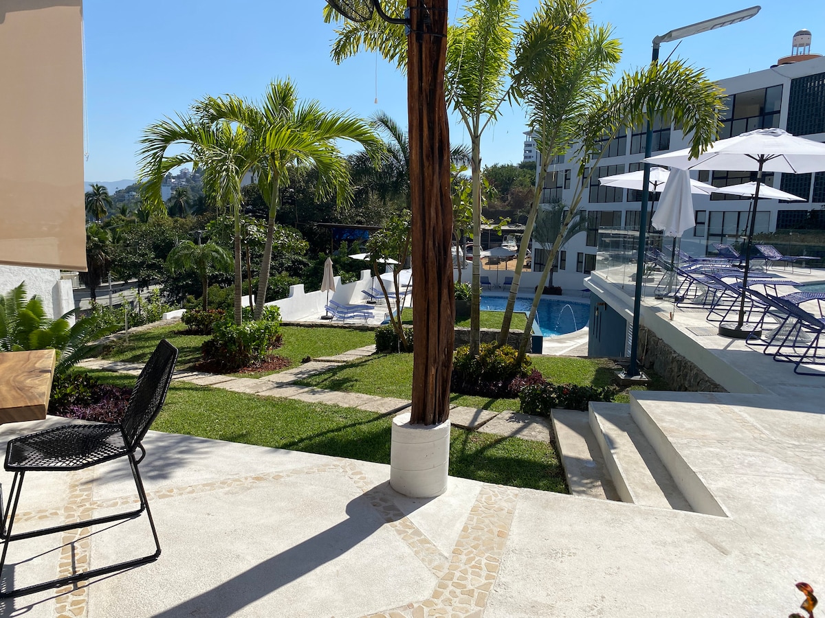 A landscaped outdoor area features palm trees and vibrant greenery. Spacious terraces and a pool area showcase lounge chairs under umbrellas. A smooth pathway leads through the garden, with a stylish black chair positioned near a potted plant, inviting relaxation and enjoyment of the scenery.