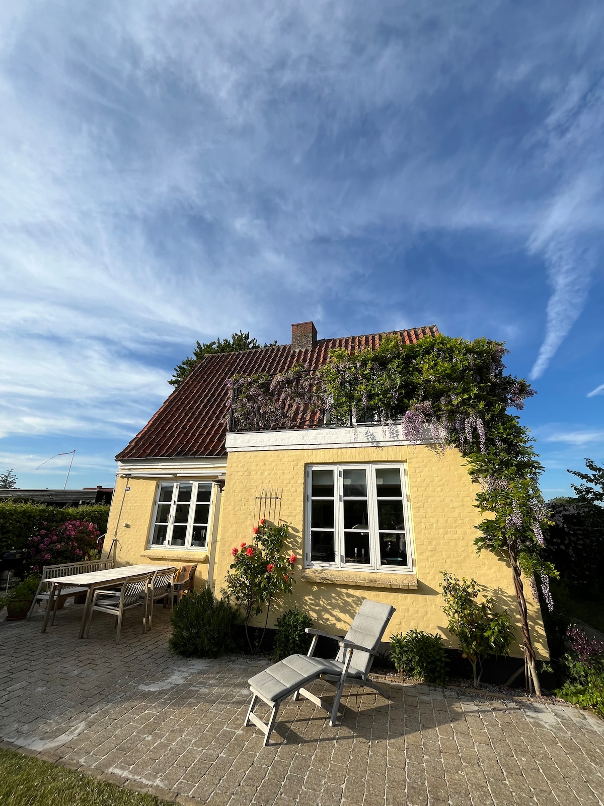 A yellow house is surrounded by greenery and colorful flowers. A wooden table and chairs are positioned in the outdoor space, ideal for enjoying the fresh air. The windows are framed with climbing plants, and a clear blue sky can be seen overhead.