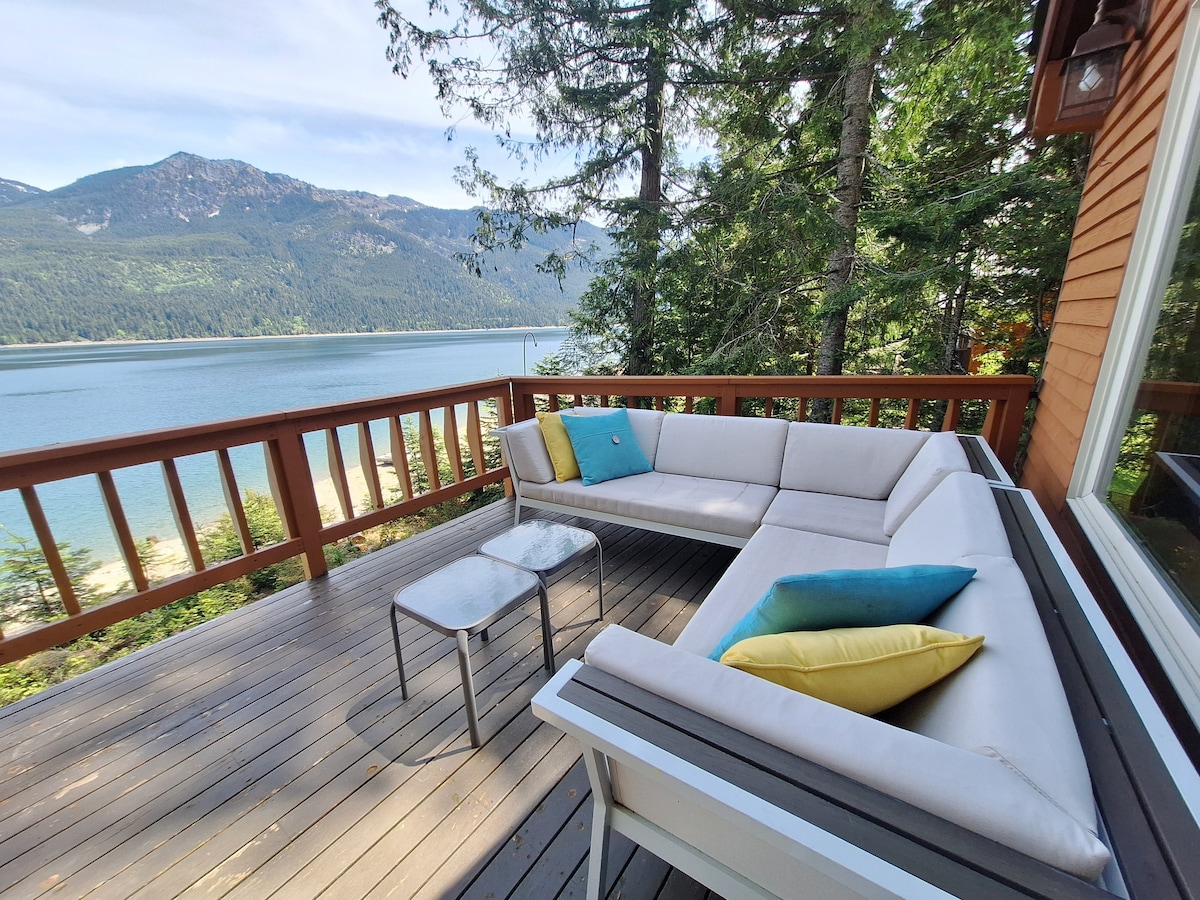A comfortable outdoor seating area is situated on a deck overlooking Lake Kachess, featuring a sectional sofa with colorful cushions and a small table. Trees frame the view of the water and mountains in the distance, creating a serene environment.
