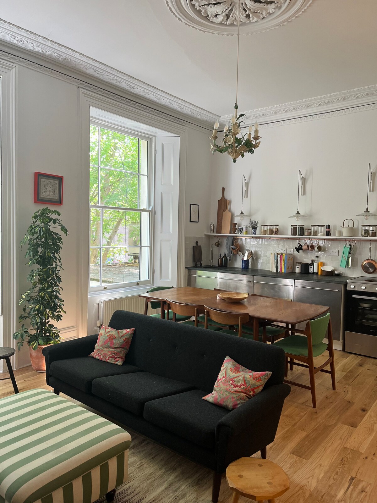 A stylish living space features a dark sofa adorned with patterned cushions. A small wooden table and green dining chairs complement the modern kitchen area, which includes stainless steel appliances and shelving. Natural light flows in through a large sash window, enhancing the room's openness.