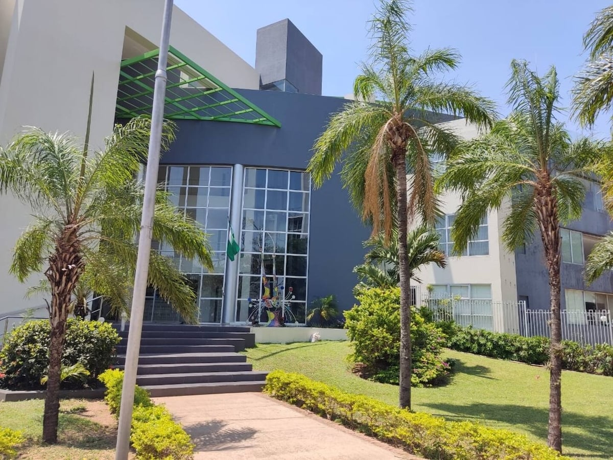 The exterior of a modern building is shown, featuring large glass windows and a prominent entrance with steps. Lush greenery and palm trees are visible in the landscaped area, enhancing the contemporary architectural design.