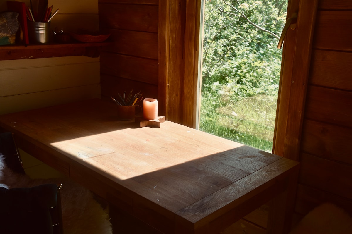 A wooden table is illuminated by natural light streaming through the open door. A candle sits on the tabletop, surrounded by art supplies. The view outside features lush greenery, creating a serene connection between the indoor and outdoor spaces.