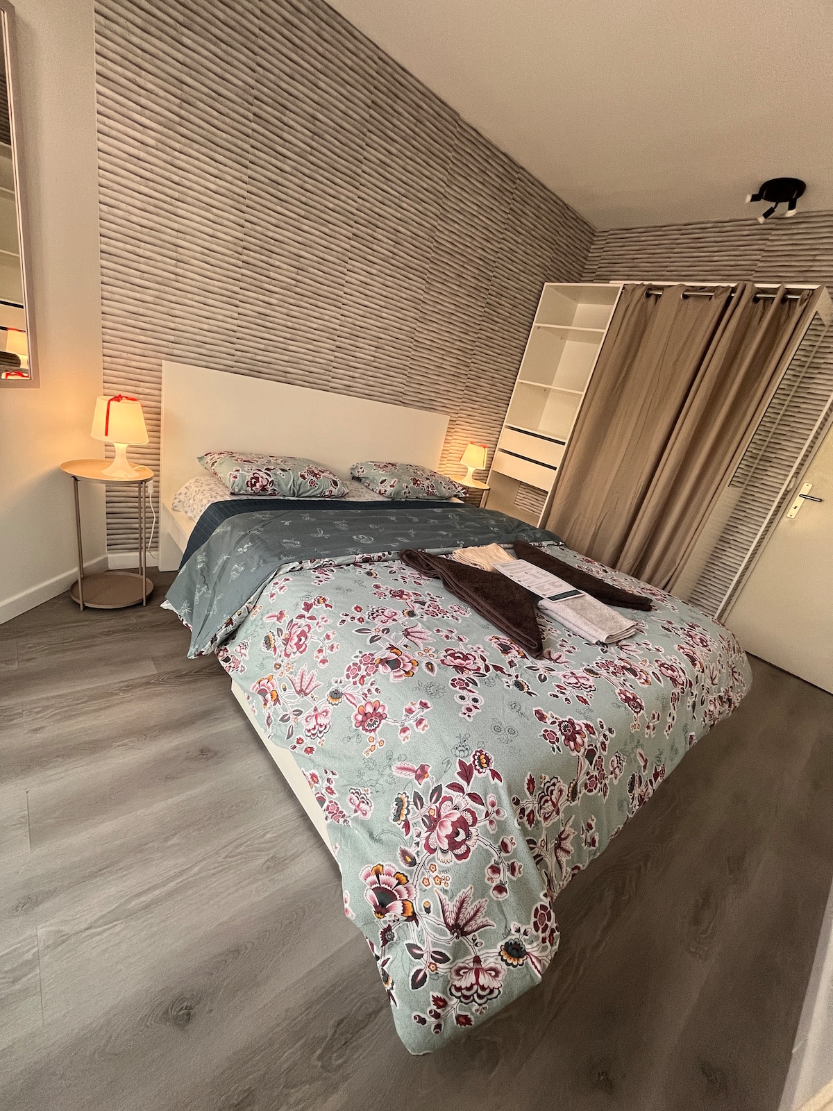A well-lit bedroom features a spacious bed adorned with floral-patterned bedding. Soft lighting is provided by two bedside lamps on circular tables. A decorative throw and folded linens are arranged neatly atop the bedding, while a wardrobe stands in the background alongside sheer curtains.