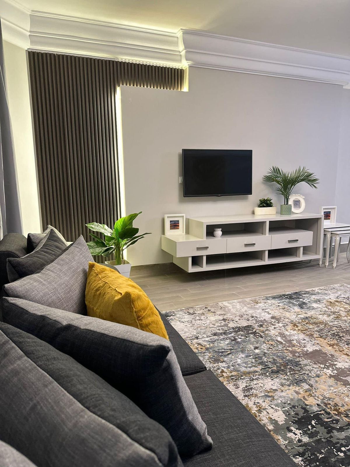 A modern living area features a plush gray sofa adorned with decorative yellow and gray cushions. Bright green plants are placed beside a minimalist entertainment unit, which supports a flat-screen television. A textured area rug adds warmth to the space, complemented by soft lighting.