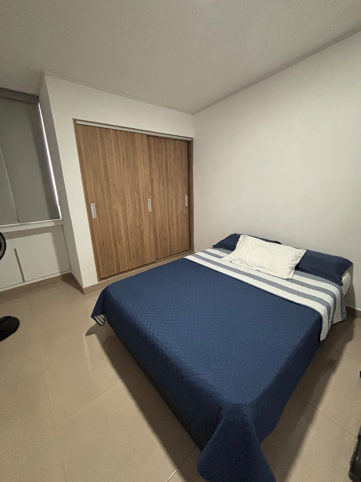 A sleeping area features a comfortable bed with a dark blue cover and a white pillow accented by a striped throw. A spacious wardrobe with wood-paneled sliding doors contrasts with the light-colored walls. Natural light enters through the nearby window.