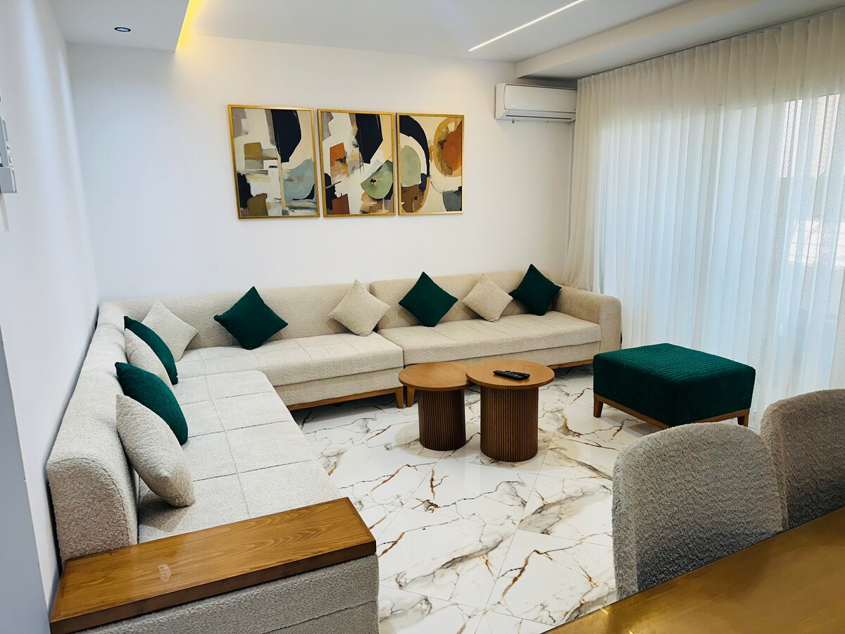 A spacious living area is presented, featuring a large L-shaped sofa adorned with green accent cushions. Two wooden coffee tables are positioned in front of the seating arrangement. Natural light filters through sheer curtains, illuminating the stylish marble flooring.