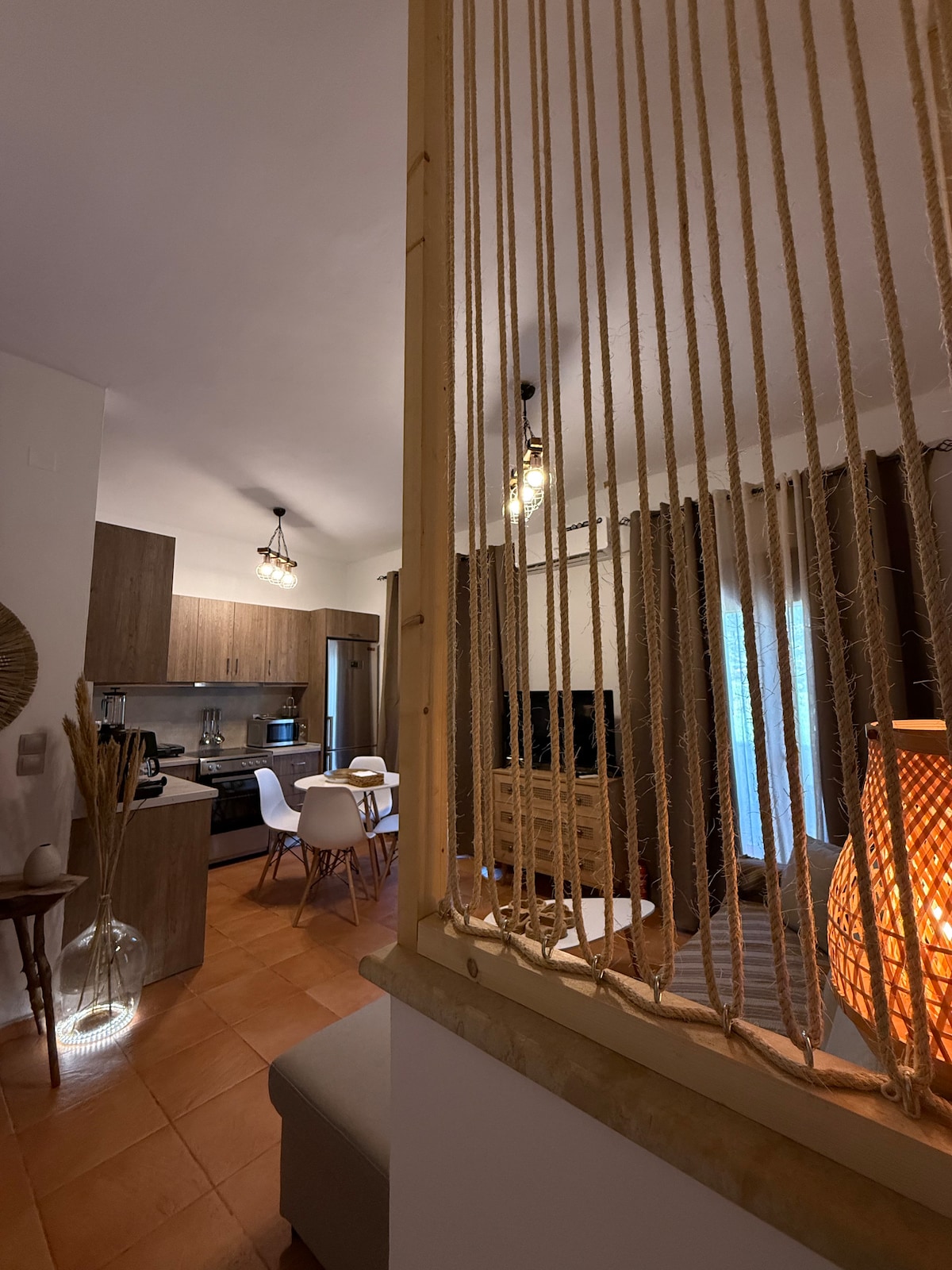 A modern living space is visible, featuring a kitchen and dining area with wooden cabinetry and a small dining table. A cozy seating area includes a television, set against a backdrop of light curtains that allow natural light to filter in, creating a warm ambiance.