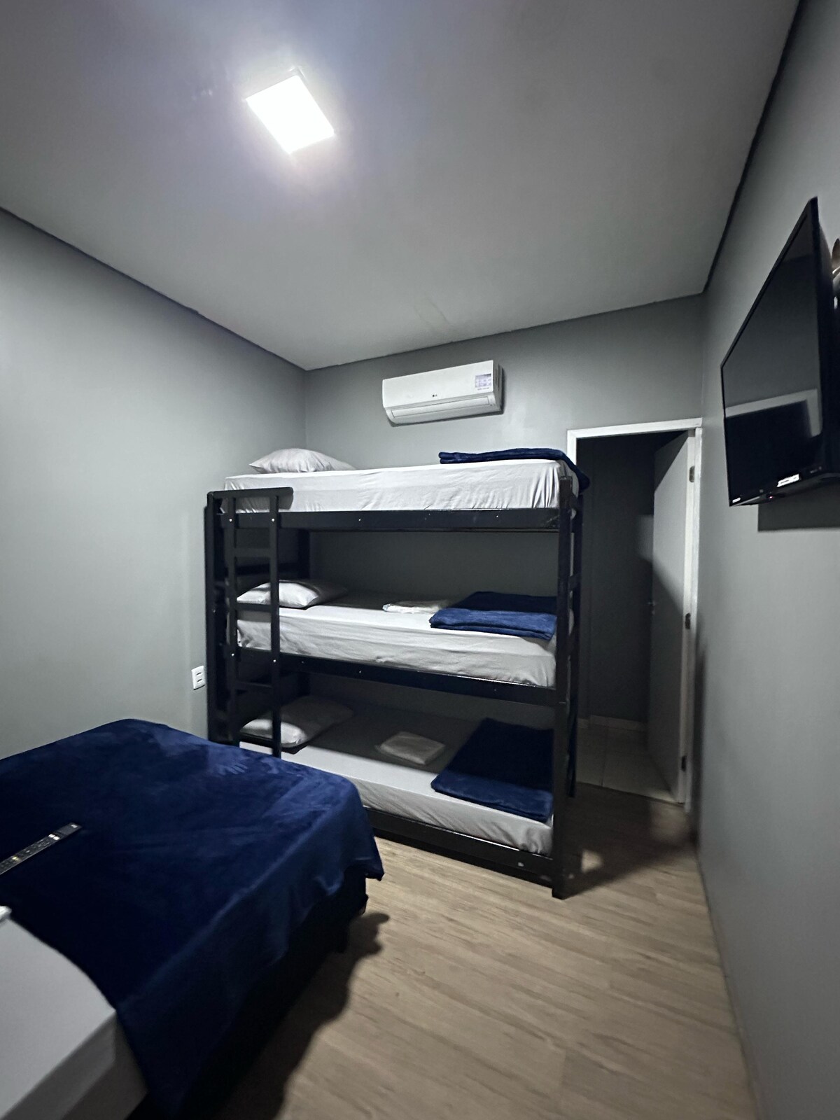 A comfortable sleeping area is arranged with a double bed and a sturdy bunk bed, featuring an upper and lower sleeping space. A flat-screen television is mounted on the wall, and a door leads to another room. Soft blue bedding adds a calming touch.