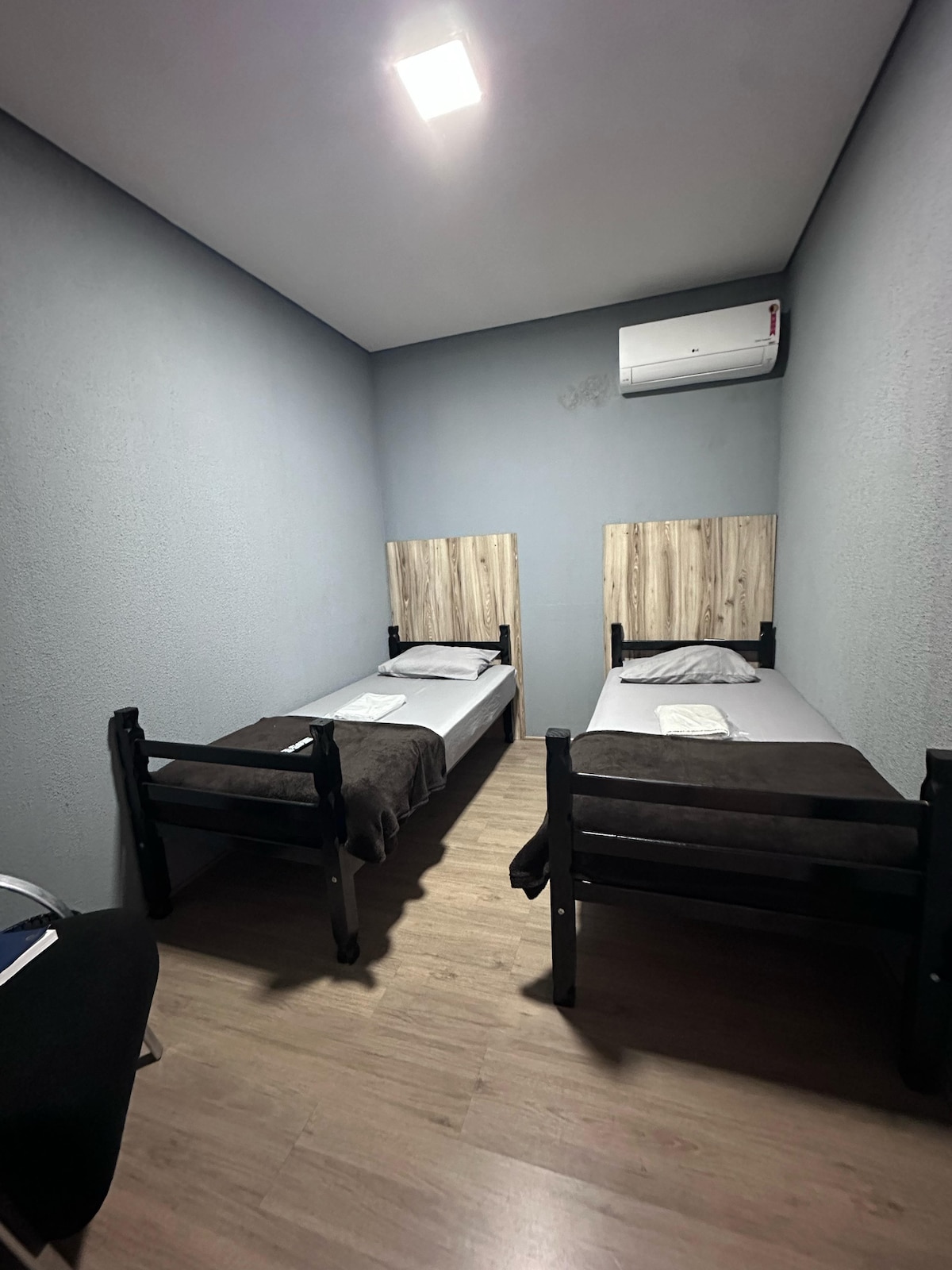 A tranquil sleeping area features two single beds, each complemented by natural wood headboards. A light-colored sheet and a dark blanket are arranged neatly on the beds. The room is painted in soft gray, with an air conditioning unit mounted above, enhancing comfort.