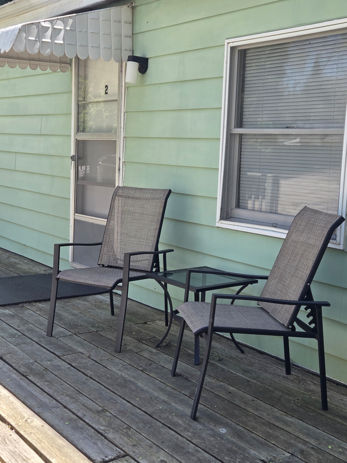 Two patio chairs are positioned next to a small table on a wooden deck. The exterior wall is painted in a soft green hue, and a window with blinds provides natural light. An awning extends above, offering shade.