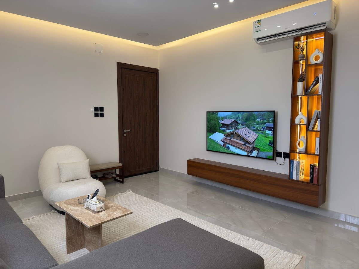 A modern living space features a comfortable lounge area with a sleek, dark sofa and a light-colored accent chair. A wall-mounted television is positioned above a wooden media console, complemented by decorative shelving with various items. Soft lighting adds a warm ambiance to the room.