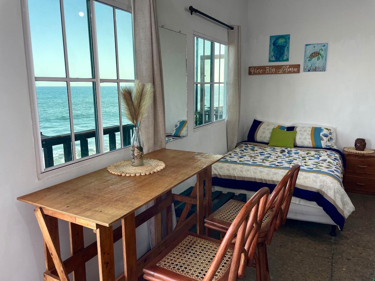 A bright and airy room showcases a spacious bed adorned with a floral duvet cover, complemented by colorful accent pillows. A wooden dining table with three chairs sits near large windows, providing unobstructed views of the ocean beyond. Art pieces and a mirror adorn the walls.