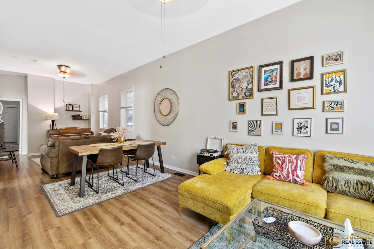 An inviting living area features a yellow sectional sofa accompanied by a glass coffee table. A large dining table with wooden chairs is visible in the background. The walls are adorned with a mix of framed artwork, creating a casual ambiance.
