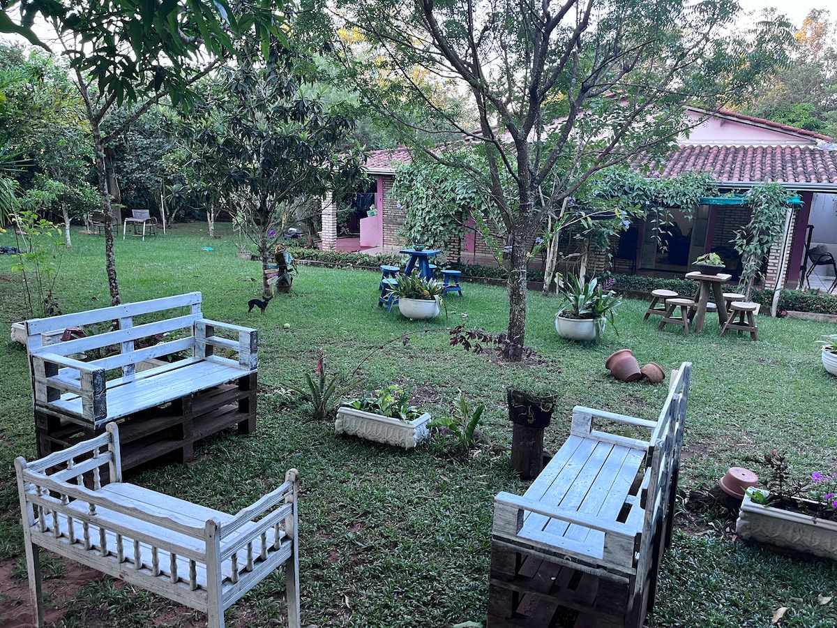 A spacious outdoor area features wooden seating arranged around a green lawn. Potted plants are placed throughout, adding natural elements. A pink building is visible in the background, surrounded by trees and greenery. The setting provides a peaceful and inviting atmosphere for relaxation.