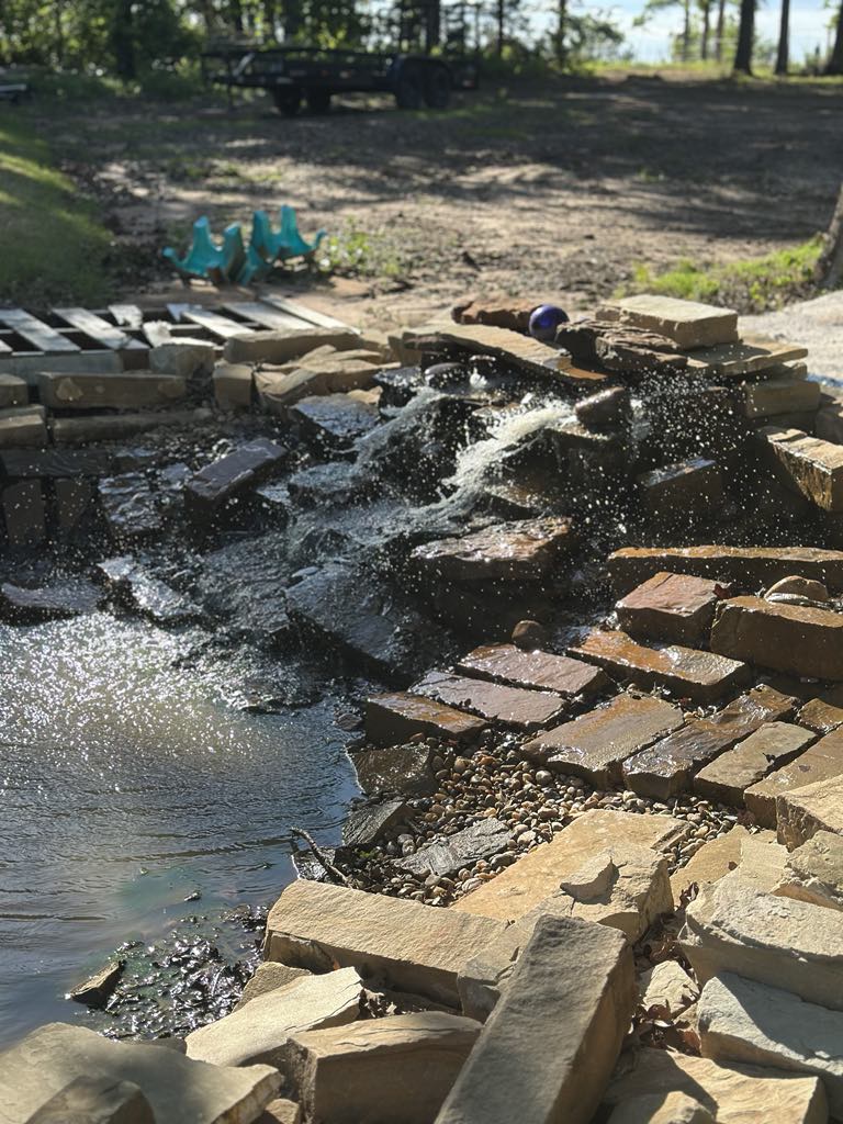 A natural stone water feature flows gently into a serene pond, creating a peaceful ambiance. Sparkling water cascades over smooth stones, while the surrounding area includes a picturesque landscape that complements the tranquil setting.