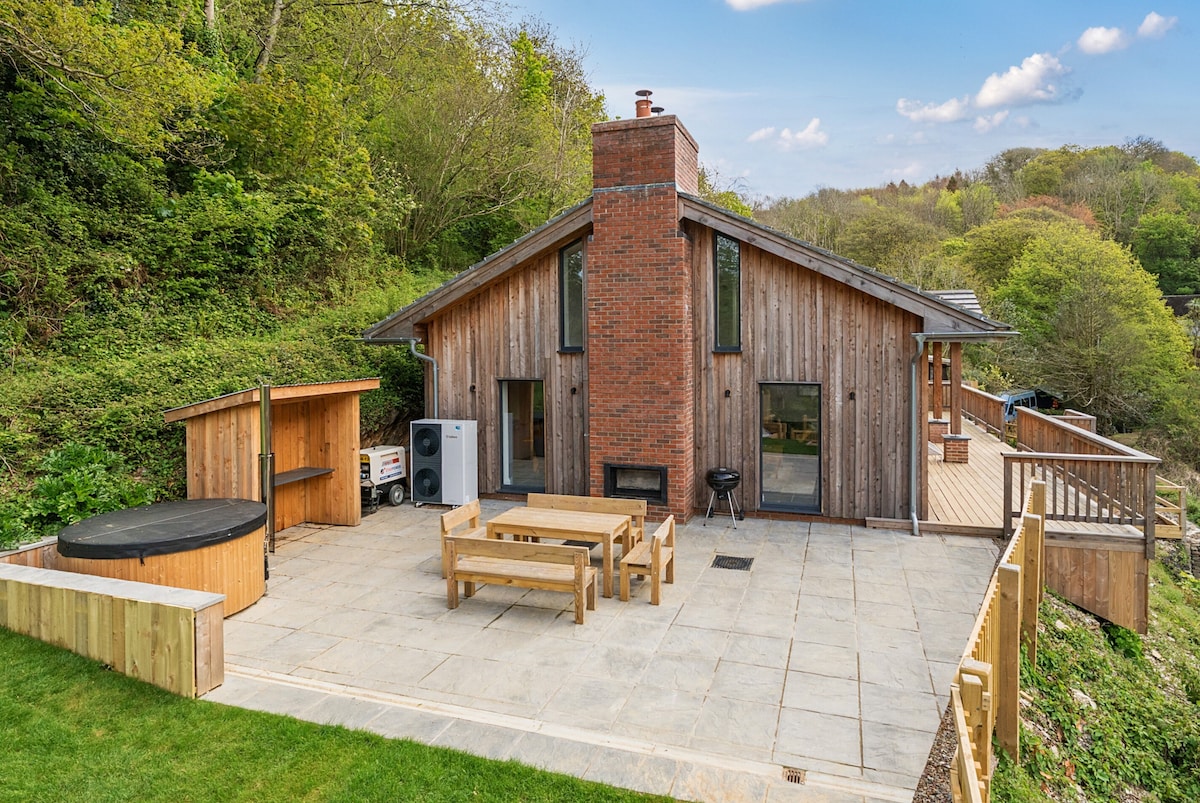 The exterior of the wooden retreat showcases a spacious patio area with outdoor dining furniture and a wood-fired hot tub. The structure features a blend of wood and brick design, set against lush greenery, with large windows that invite natural light.