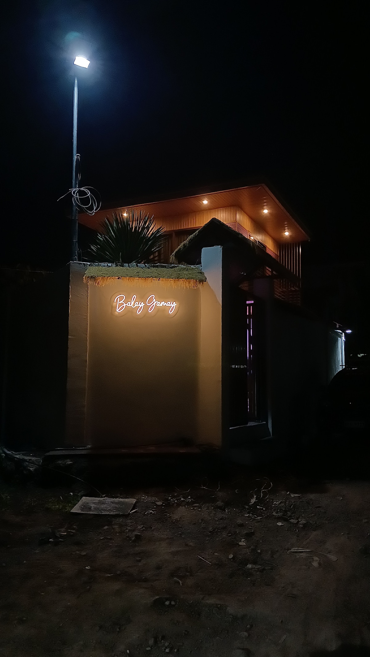 The exterior of the property is illuminated by a soft light, highlighting the modern architectural features. A neon sign displaying the name 'Balay Gamay' is mounted on a wall, creating a welcoming visibility at night. Lush greenery frames the entrance.
