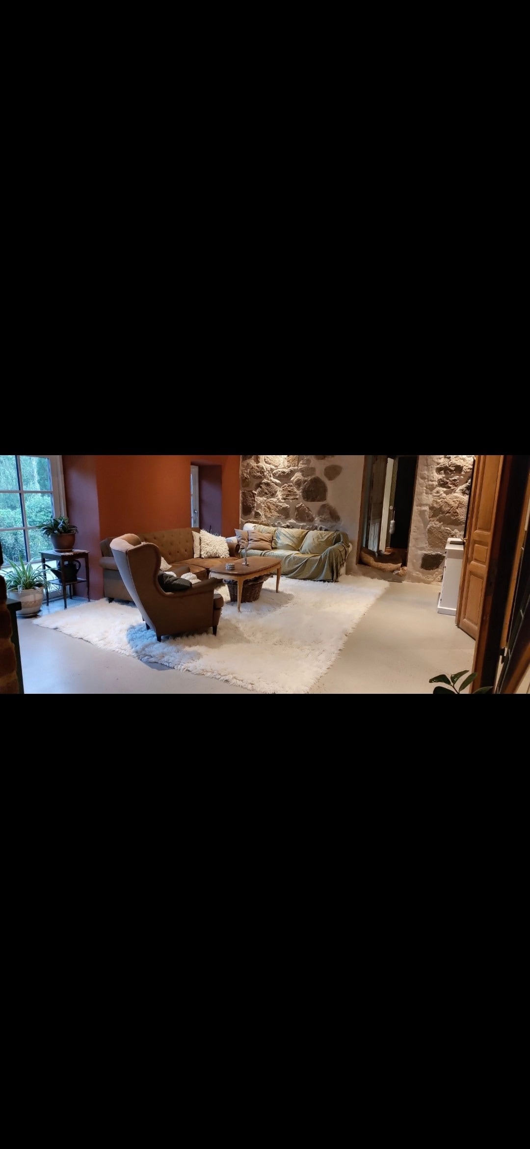 A cozy sitting area is present, featuring a comfortable armchair and a small table placed on a soft rug. Stone walls provide a rustic backdrop, while a window frames an outdoor view. Textiles in neutral tones add warmth to the space.