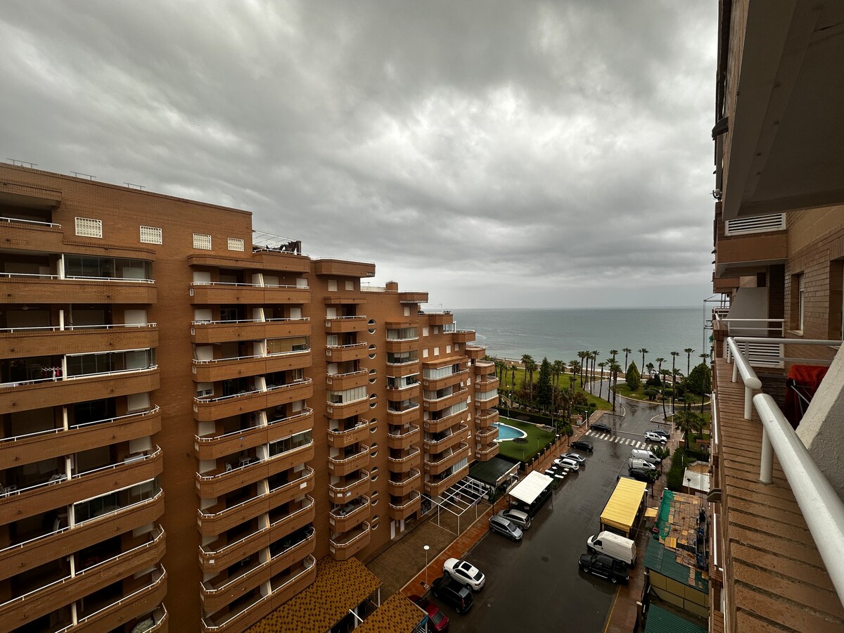 An expansive view showcases residential buildings along a coastal area, with the ocean visible in the distance. A cloudy sky blankets the scene, while palm trees and a swimming pool are seen below, complementing the surroundings.