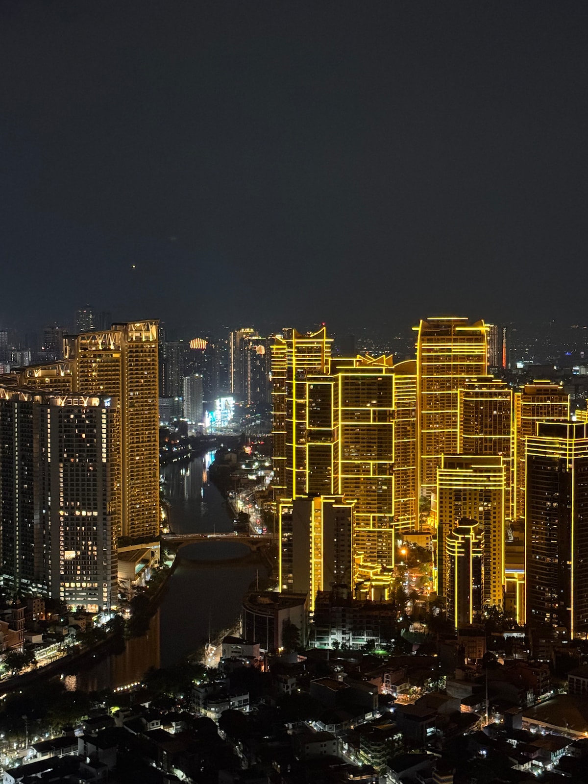The city skyline is illuminated at night, showcasing tall buildings adorned with bright yellow lighting. A river runs through the foreground, reflecting the vibrant lights of the urban landscape. The view captures both modern architecture and a lively atmosphere.