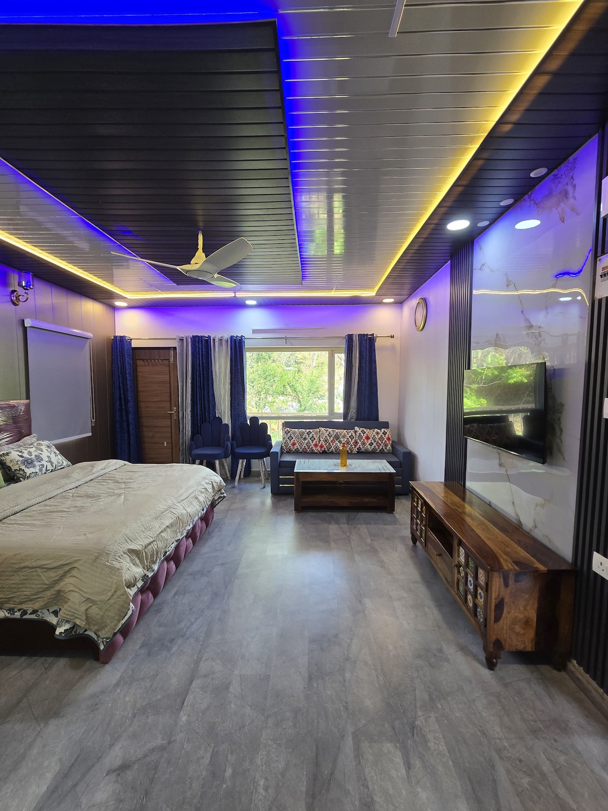 A cozy studio apartment features a spacious bed adorned with neutral bedding. A comfortable sofa and coffee table create a relaxed seating area, complemented by large windows that allow natural light to brighten the room. Air conditioning and wall-mounted TV enhance the modern interior.