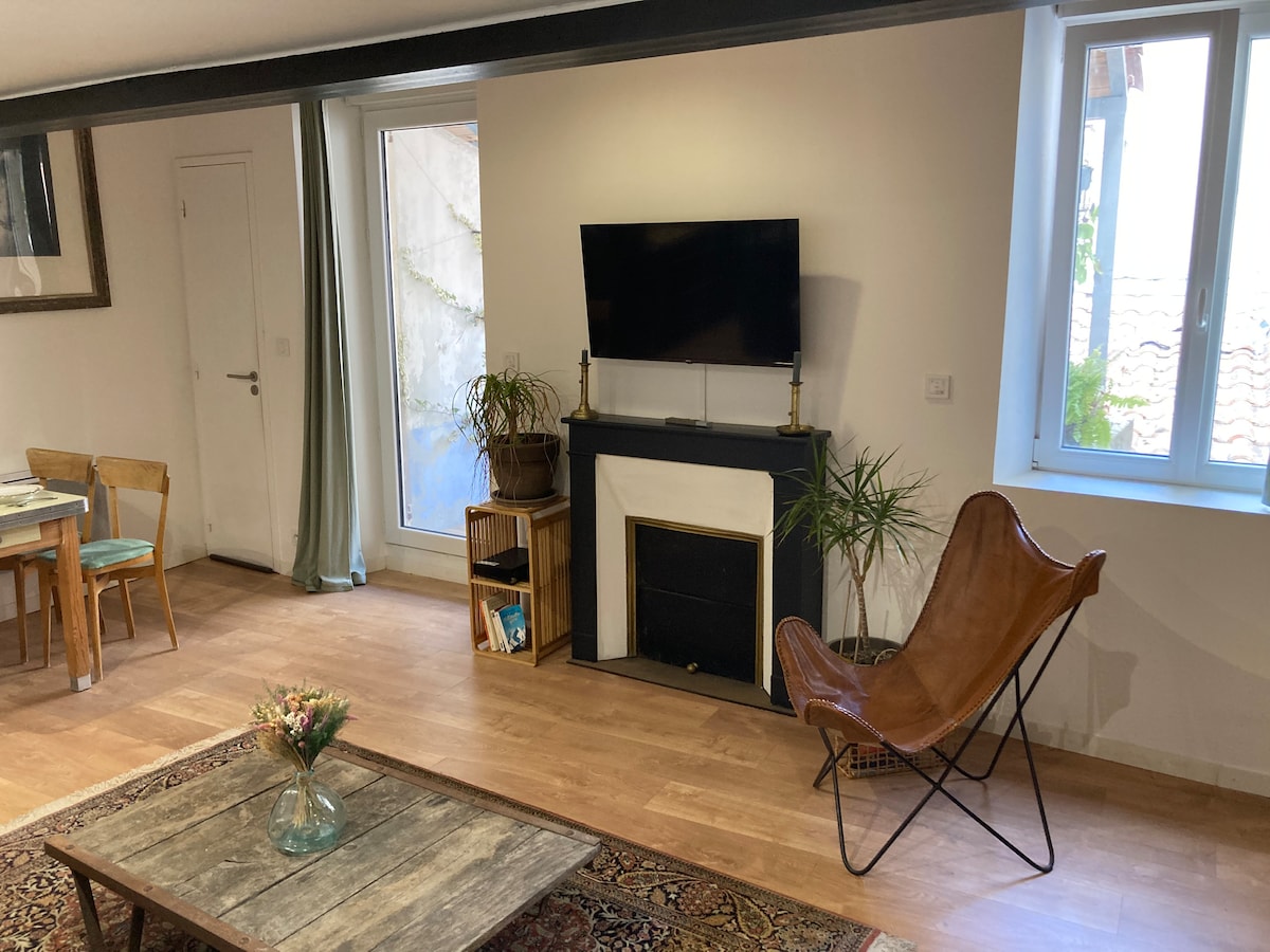 A living area is shown with warm wood flooring, featuring a comfortable leather chair and a simple coffee table. A flat-screen television is mounted above a black fireplace. Natural light enters through two large windows, and a small indoor plant adds a touch of greenery.