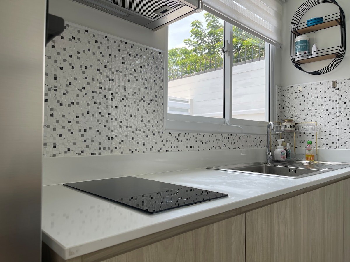 A modern kitchen displays sleek countertops with a built-in cooktop, complemented by a stainless steel sink. Mosaic tiles in black and white adorn the walls, while natural light floods in through a window overlooking a garden area.