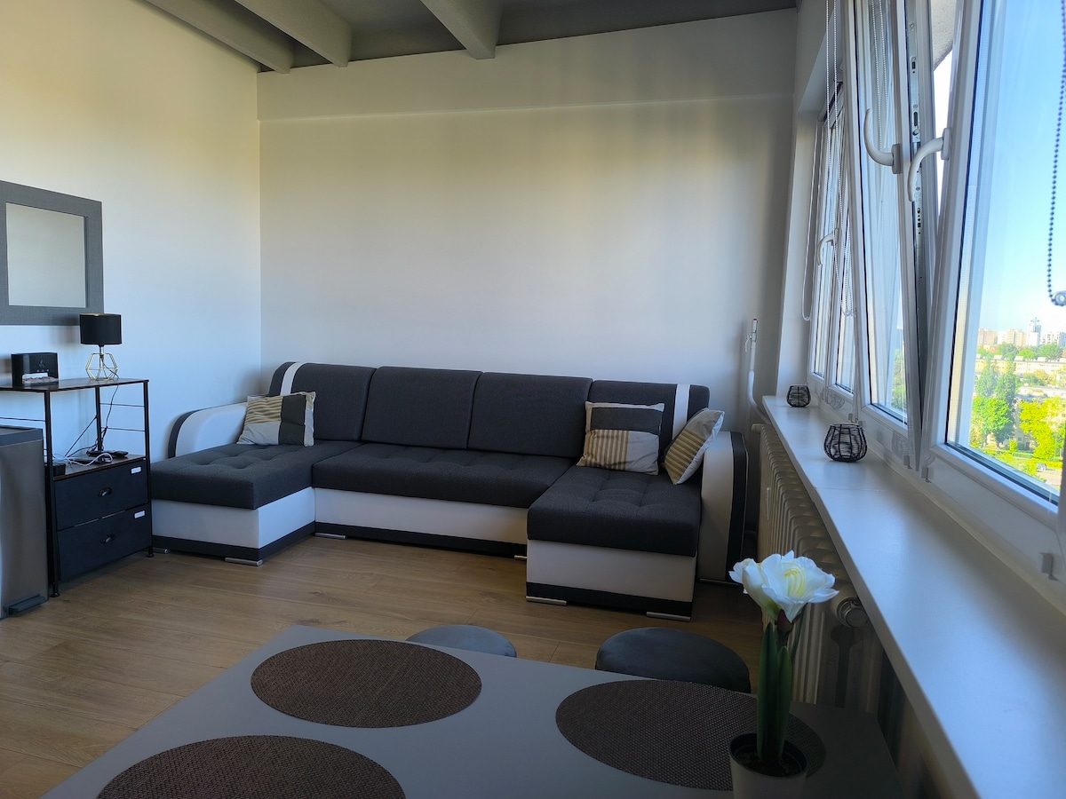 The living area features a modern sectional sofa in black and white, complemented by decorative cushions. A sleek coffee table with circular placemats sits in front of the sofa. Large windows offer a view of the surroundings, allowing natural light to fill the space.