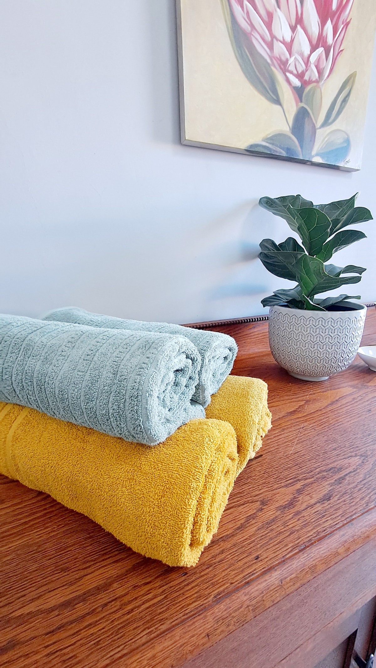 A wooden surface is adorned with neatly folded towels in soft blue and vibrant yellow. A potted plant with lush green leaves adds a touch of freshness. A floral artwork in muted tones hangs on the wall, enhancing the serene setting.