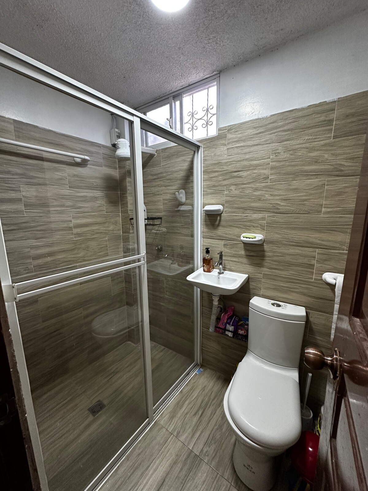 A clean and functional bathroom is presented, featuring a glass shower enclosure and a modern toilet. Light-colored tiles cover the walls and floor, while a small shelf holds toiletries. A window above the shower allows natural light to enter the space.