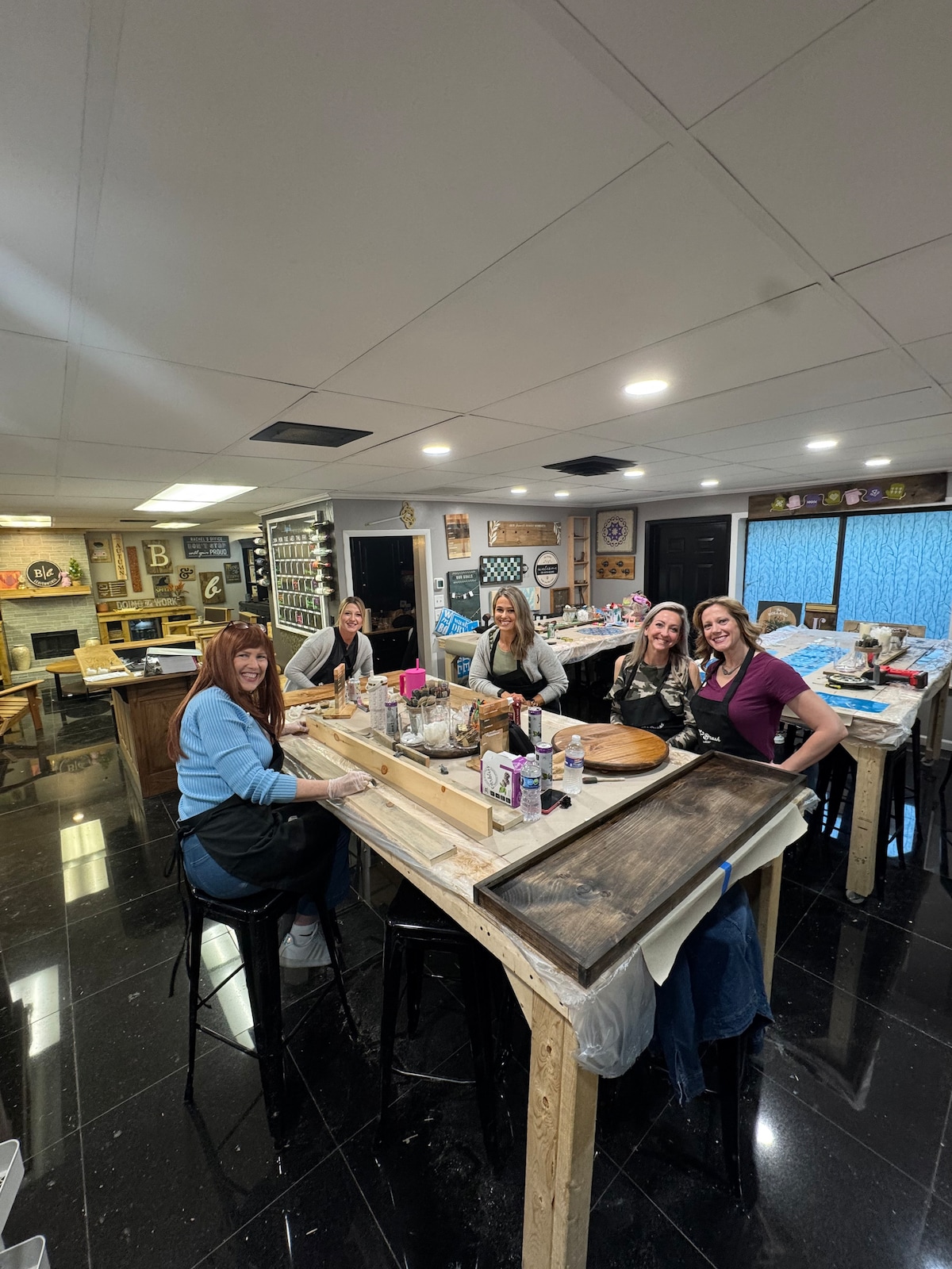 A well-lit workshop space is shown with several individuals focused on crafting projects at a long wooden table. The floor features polished black tiles, while various art supplies and tools are neatly organized in the background, showcasing a creative atmosphere.