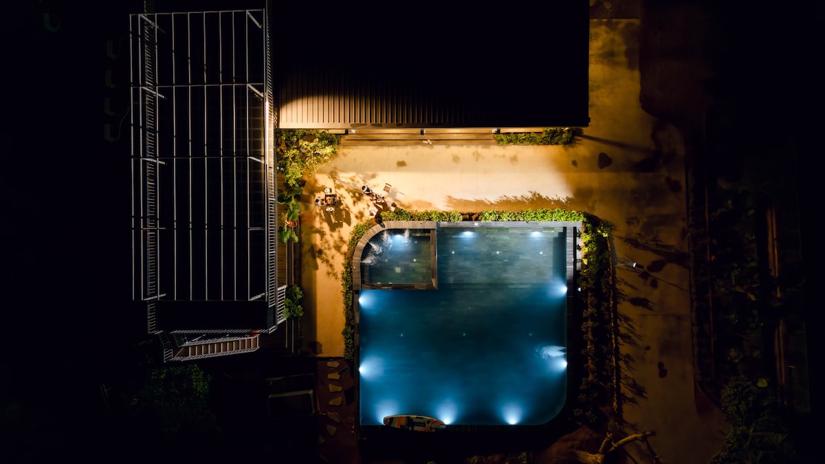 An aerial view captures a tranquil pool surrounded by lush greenery and illuminated by soft lighting. The pool is bordered by a pathway, and the nearby structure features a sloped roof, enhancing the serene outdoor environment.